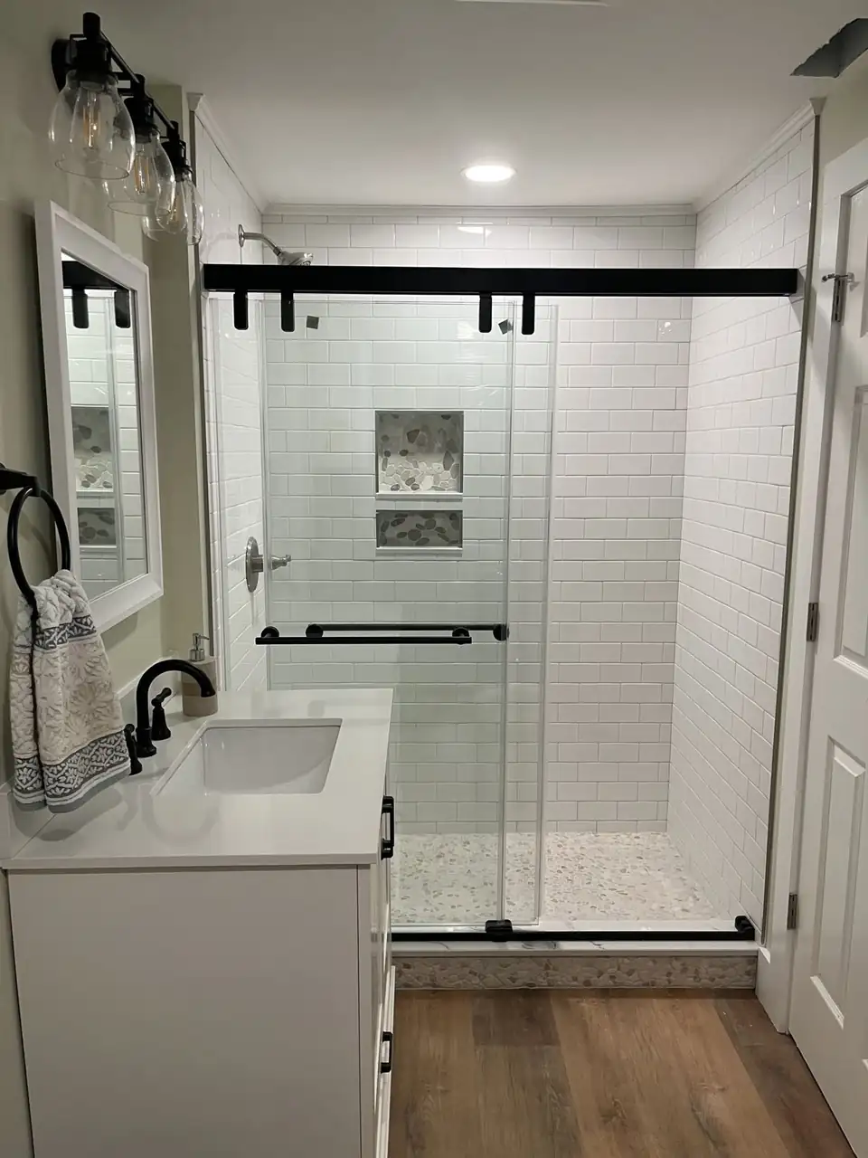 Bathroom remodeling in Buffalo by CNG Construction: white subway tile shower with a pebble floor and mosaic niche, black sliding glass door and white vanity