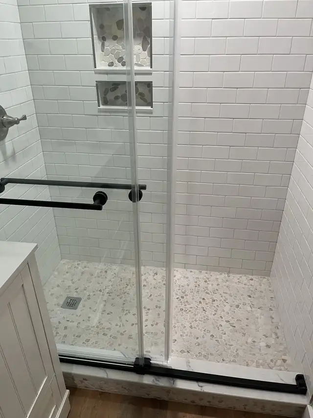 White subway tile shower with a pebble floor and black hardware on the glass door, from a full cottage renovation